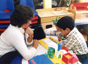 A photograph shows a woman seated at a table across from a young male child. The woman has short, dark hair and is wearing a white turtleneck. The child is of primary school age, and is concentrating on a puzzle while the woman watches.
