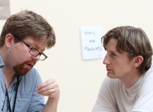 A photograph shows a man speaking while another man listens. The man at left is the speaker; he is looking down at a point outside the lower edge of the frame and using his left hand to gesticulate. The man at right is watching the speaker’s face.