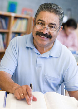 A photograph shows a middle-aged man smiling at the camera. He has a dark moustache and wears glasses. The man is holding a pen over an open book on a surface in front of him; he appears to be in the act of underlining or pointing to text. Bookshelves are visible in the background.