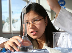 A photograph shows a young woman in a lab coat and glasses using a pipette to transfer a clear liquid from a beaker into a volumetric flask. The woman is crouching beside the lab bench so that she is eye-level with the meniscus of the flask. She is using her right hand to support the flask, and her left hand to operate the pipette.