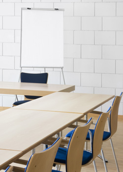 A photograph depicts an empty classroom space. Three tables with chairs behind them are perpendicular to a fourth table at the front of the classroom. Behind the fourth table is an empty chair and a large blank notepad on an easel.
