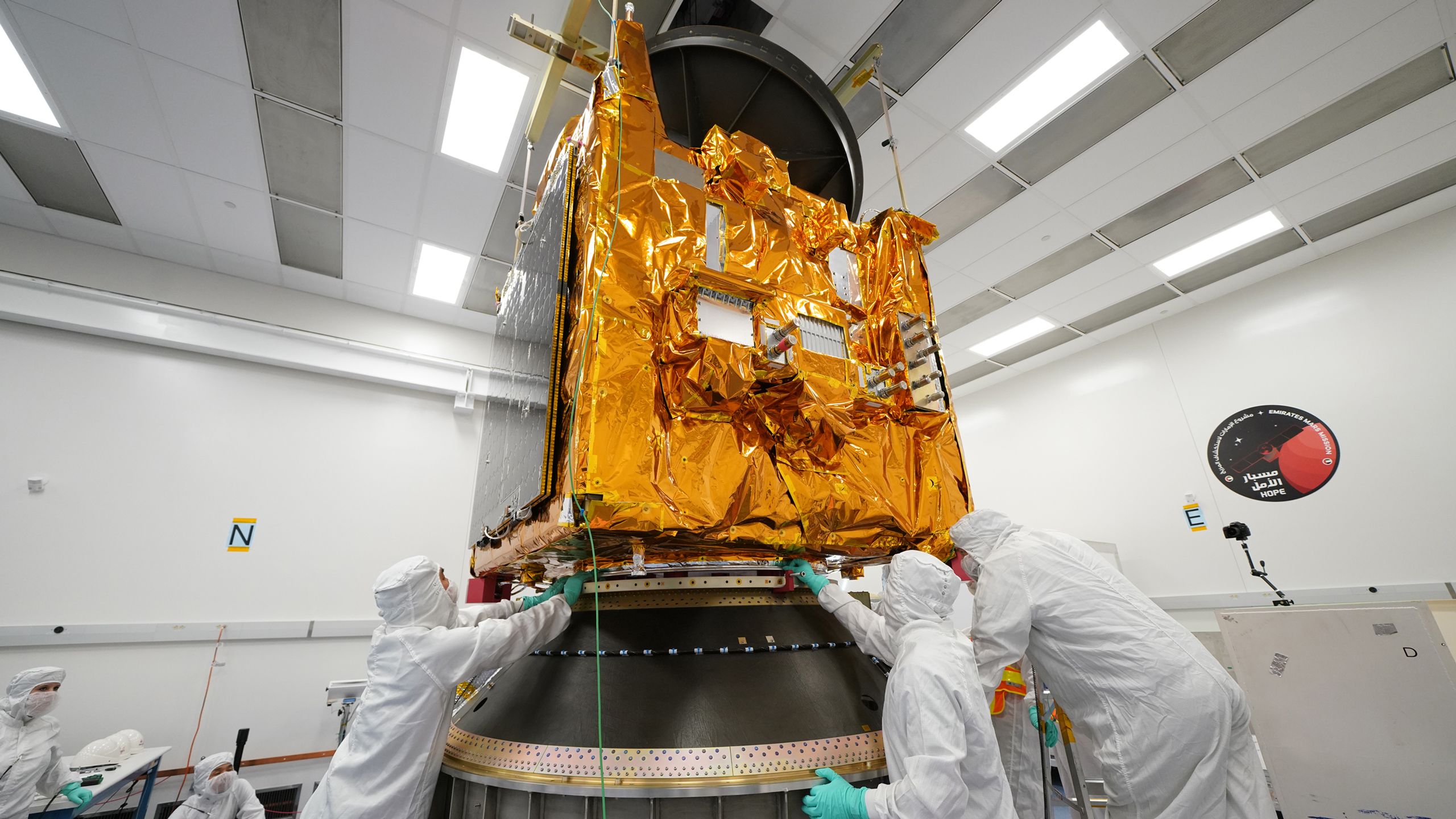 Engineers in white clean room suits reaching up towards the gold coloured central section of the Hope Probe.