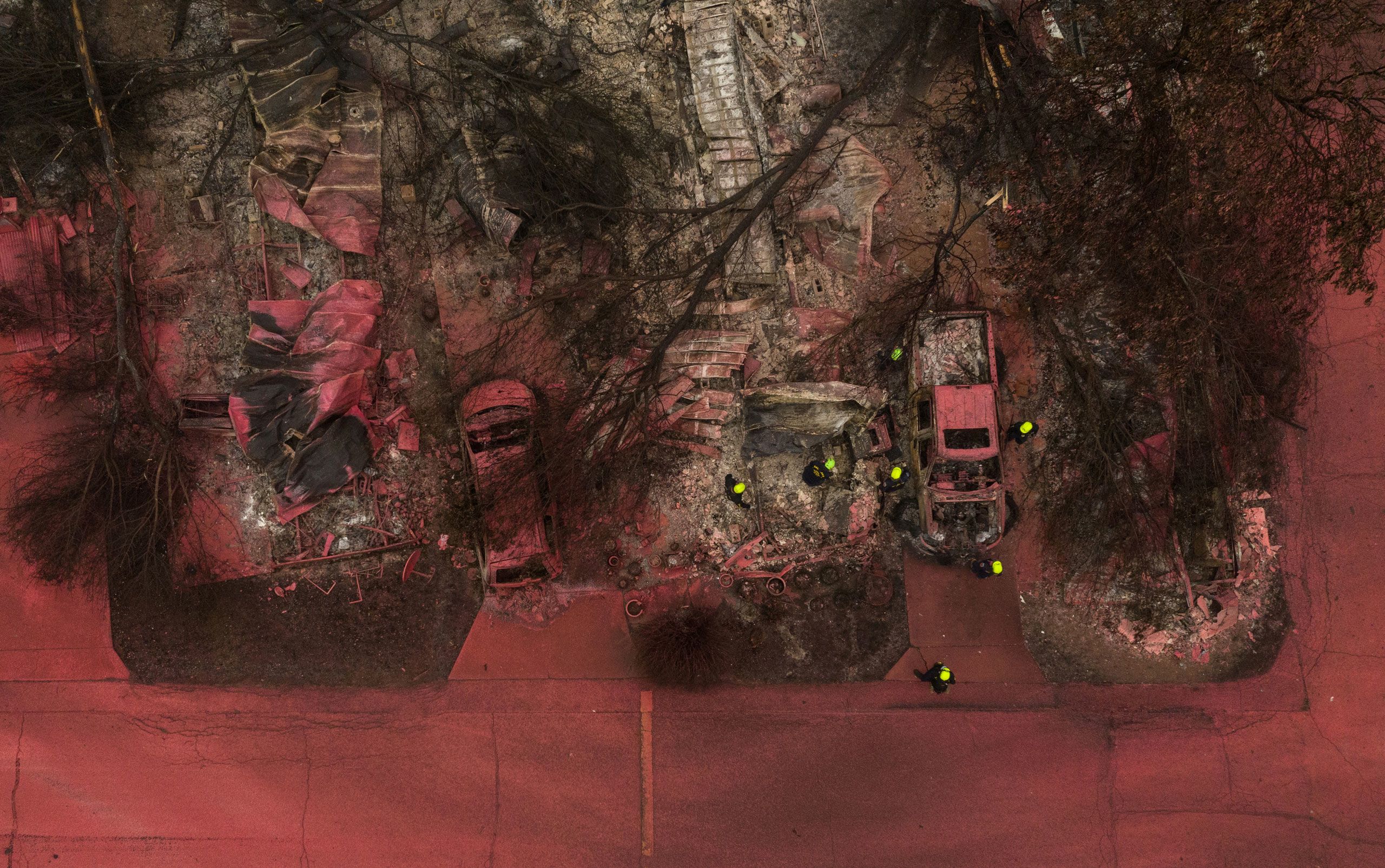 A search and rescue team, surrounded by red fire retardant, look for victims under burned residences and vehicles.