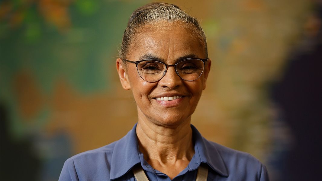 Portrait Marina Silva wearing a blue shirt and glasses, with hair tied back in front of a blurred background.