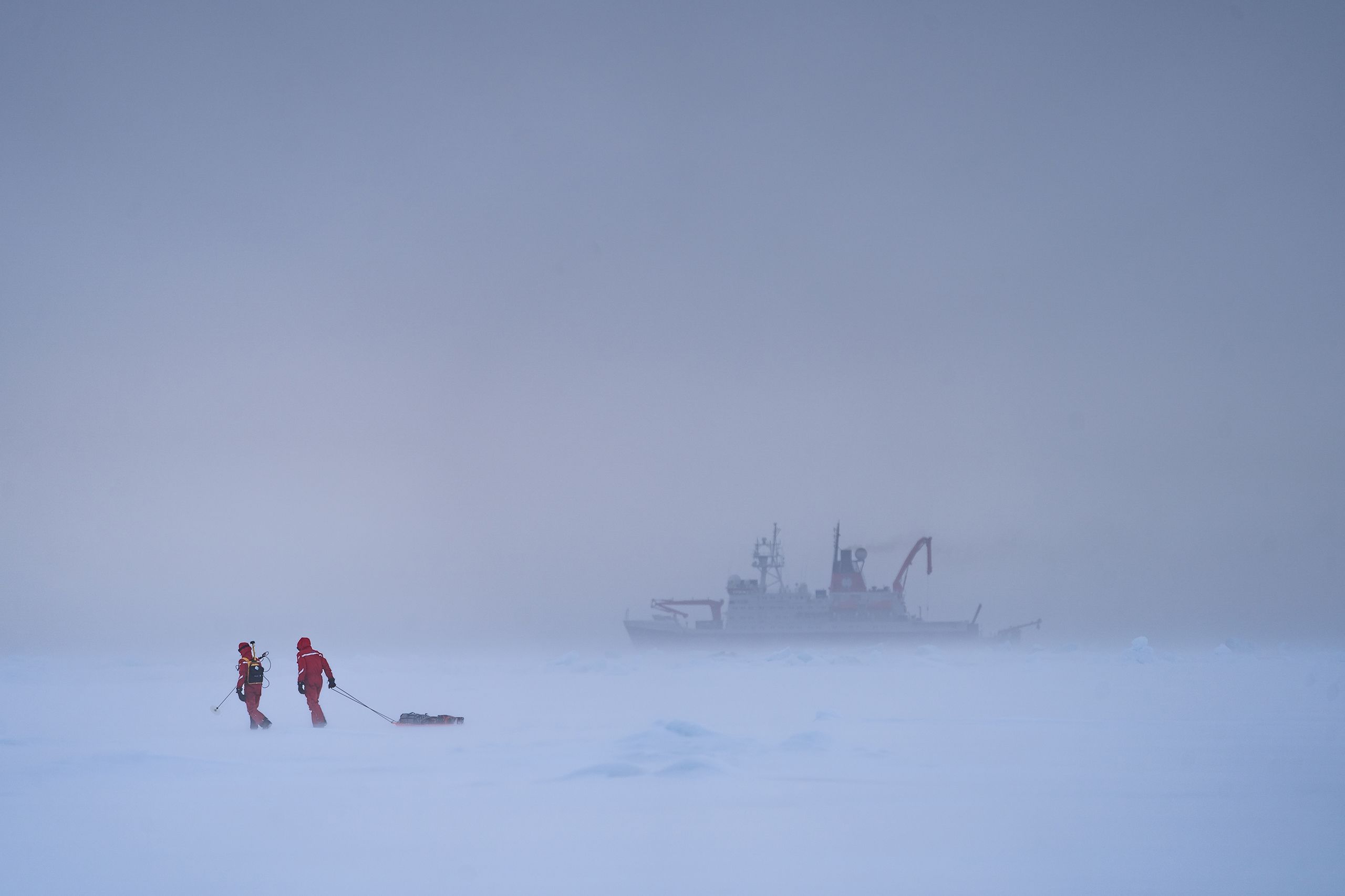 Two scientists drag equipment along on a sled over an ice floe while the Polarstern research vessel is visible through mist in the distance