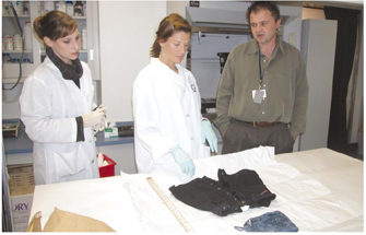 A photograph of a crime laboratory shows two women in white lab coats and latex gloves standing beside a man in an olive green shirt and grey slacks. In front of them, clothing items are arranged on a table beside a wooden ruler. The woman at center is reaching towards the clothing with her right hand while the other two watch.