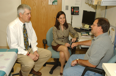 A photograph shows a woman sitting between two men seated on chairs in a medical office. The woman and one man are dressed in business attire and are facing the second man, who is dressed in a grey-collared shirt and blue jeans. The woman is smiling and holding a pen.