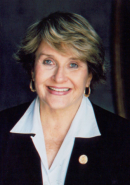 A photograph shows a front-facing portrait of a woman, Representative Louise Slaughter, smiling at the camera. She is wearing a white collared blouse under a dark suit jacket. Representative Slaughter’s dark blonde hair is cut above her ears. She is wearing gold hoop earrings and a gold button on the left lapel of her jacket.