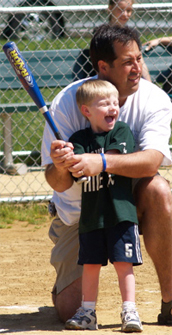 A photograph shows a young boy standing at home plate on a baseball diamond holding an aluminum bat and laughing. The child's father kneels on one knee behind him and holds the bat in the same stance for support.