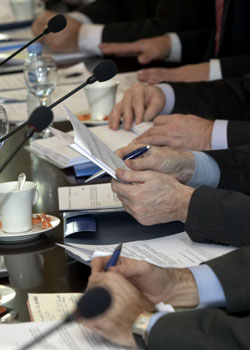 A photograph shows the hands from five individuals that are sitting in a row at a table. The five individuals are wearing black suit jackets over white or blue dress shirts. There are mugs and water glasses in random positions along the table. In front of each pair of hands are papers and microphones. The two individuals whose hands are shown in the foreground are holding pens. The second individual appears to be reading from a paper packet.