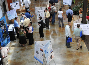 A photograph shows an aerial view of an indoor internship fair. Several poster boards are assembled on easels at various spots in the room. Several people in business-casual attire are engaged in conversation near each poster.