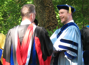 A photograph shows a professor and student engaged in conversation at a graduation ceremony. The professor and student both wear graduation gowns.