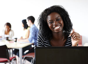 בתצלום נראית אישה צעירה מחייכת למצלמה מאחורי מחשב נייד.  ברקע, שתי סטודנטיות וסטודנט אחד יושבות יחד ליד שולחן.  השולחן מכוסה בניירת ובכוסות קפה.