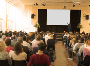 A photograph shows an audience of people sitting in rows with their backs to the camera. In front of the audience, a man stands on a stage and points at a white screen.