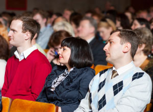A photograph shows an auditorium of people viewing a presentation. A woman and two men are in the foreground of the frame; their heads are turned in the same direction, towards the speaker.
